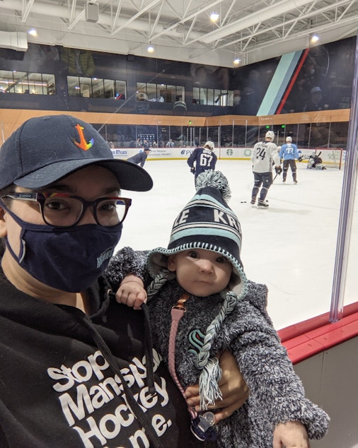 caption: Christy Maggio, a mega hockey fan, with their baby. Maggio loves the Kraken, Seattle's new hockey team, but laments misogyny heard from the stands.