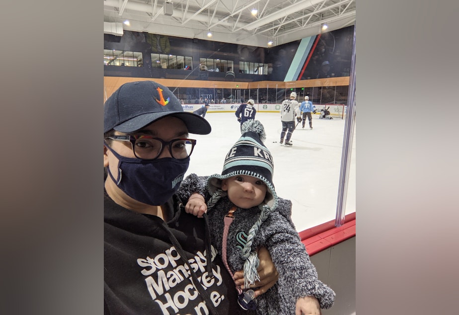 caption: Christy Maggio, a mega hockey fan, with their baby. Maggio loves the Kraken, Seattle's new hockey team, but laments misogyny heard from the stands.