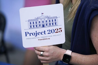 caption: A woman holds a Project 2025 fan in the group's tent at the Iowa State Fair, Aug. 14, 2023, in Des Moines, Iowa. (Charlie Neibergall/AP)