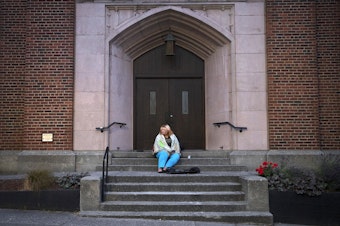 caption: Milee Ballweg sleeps on the steps of a church in Seattle's University District. She's 20, her hair is blonde tinged with pink, and she wears ripped jeans and a PokÃ©mon T-shirt. She sleeps wrapped in gray packing blankets that cover her face. 