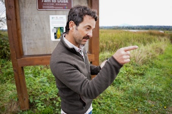 caption: Brett Shattuck, points to the different walkways in and around Spencer Island. The main asphalt road is eroding away, and pathways leading down to the water have been grown over. 