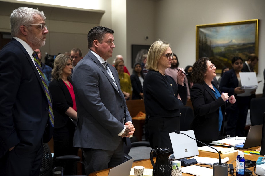 caption: Jeffrey Nelson, center, stands during his trial between his defense attorneys Tim Leary, left, and Emma Scanlan, right, as King County Superior Court Judge Nicole Gaines Phelps enters the courtroom on Wednesday, May 22, 2024, at the Maleng Regional Justice Center in Kent. 