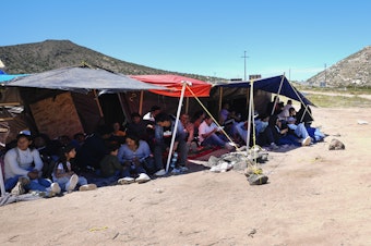 caption: A group of people wait to be processed after crossing the border between Mexico and the United States as they seek asylum in April 2024, near Jacumba, Calif.