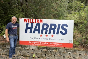 caption: William Harris, Republican candidate for Mason County Commissioner, with one of his campaign signs that he modified after Vice President Kamala Harris became the Democratic nominee.