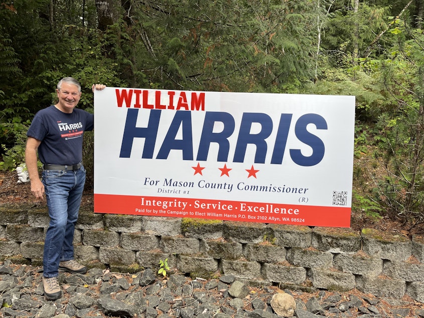 caption: William Harris, Republican candidate for Mason County Commissioner, with one of his campaign signs that he modified after Vice President Kamala Harris became the Democratic nominee.