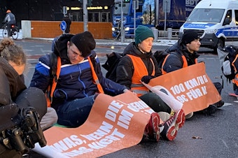 caption: Climate activists from the group Letzte Generation (Last Generation) hold up commuter traffic on a Monday morning in Berlin by supergluing themselves to the road. Police unstick their hands using cooking oil and a pastry brush while irate drivers look on, stuck for more than an hour.