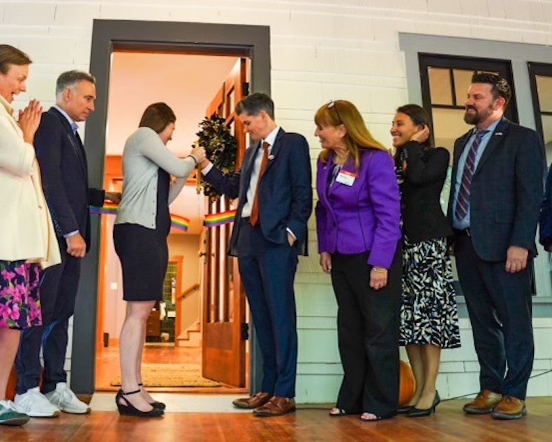 caption: Lindsay Church, in the suit on the right side of the doorway, is joined by loved ones, local, state, and federal officials marking the opening of a new transitional housing program for LGBTQ+ veterans. Also in attendance were representatives from Veterans Affairs, King County Executive Dow Constantine, and Congressmembers Pramila Jayapal and Adam Smith.