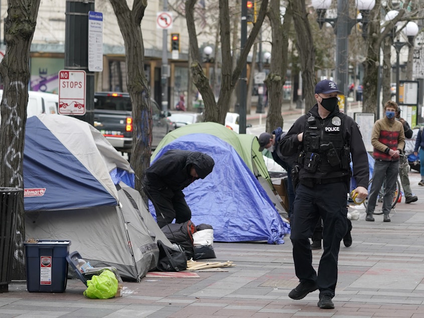 caption: A Seattle police officer walks past tents used by people experiencing homelessness, March 11, 2022, during the clearing and removal an encampment in Westlake Park in downtown Seattle.