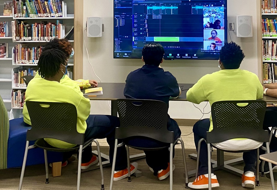 caption: Five youth sit around a table and listen as their audio stories play over a large computer screen on April 15, 2021.