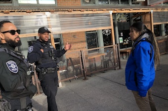 caption: Community crisis responder Sasha Pollock, right, checks in with Seattle police officers before approaching a crisis call in Seattle's Belltown neighborhood in December 2023. 