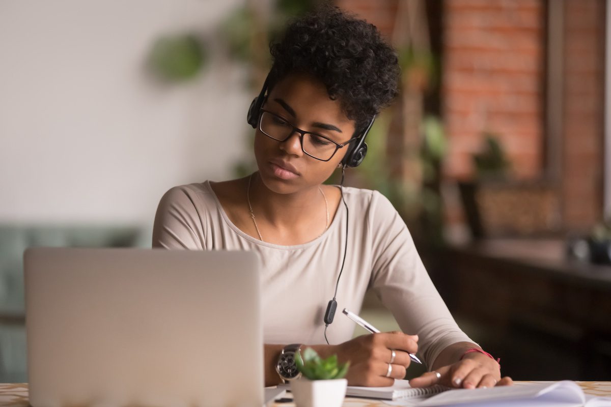 Focused mixed race woman wearing headphones watching webinar write notes study online with skype teacher, african female student learning language computer course on laptop listen translate lecture